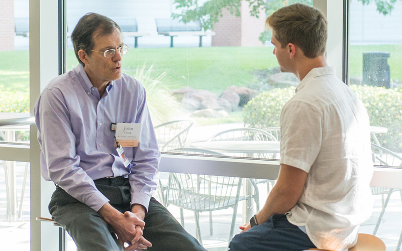 Two people, both seated on stools, one older male speaking with a younger male