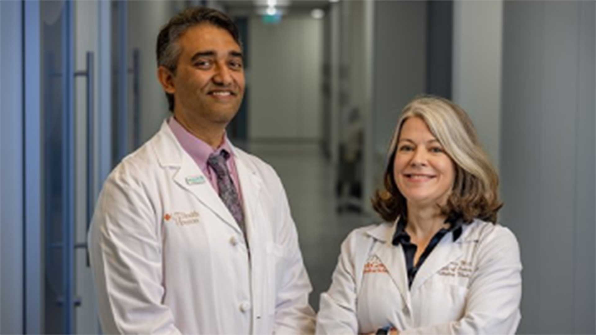Posed image of Dr. Aanand Naik, MD and Dr. Holly M. Holmes, MD in white coats standing side by side at the beginning of a long hallway