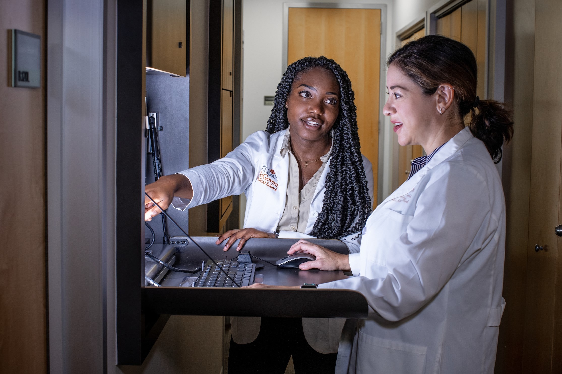 Two female doctors in white coats at a laptop terminal are caught in discussion, one of which is pointing at the laptop screen